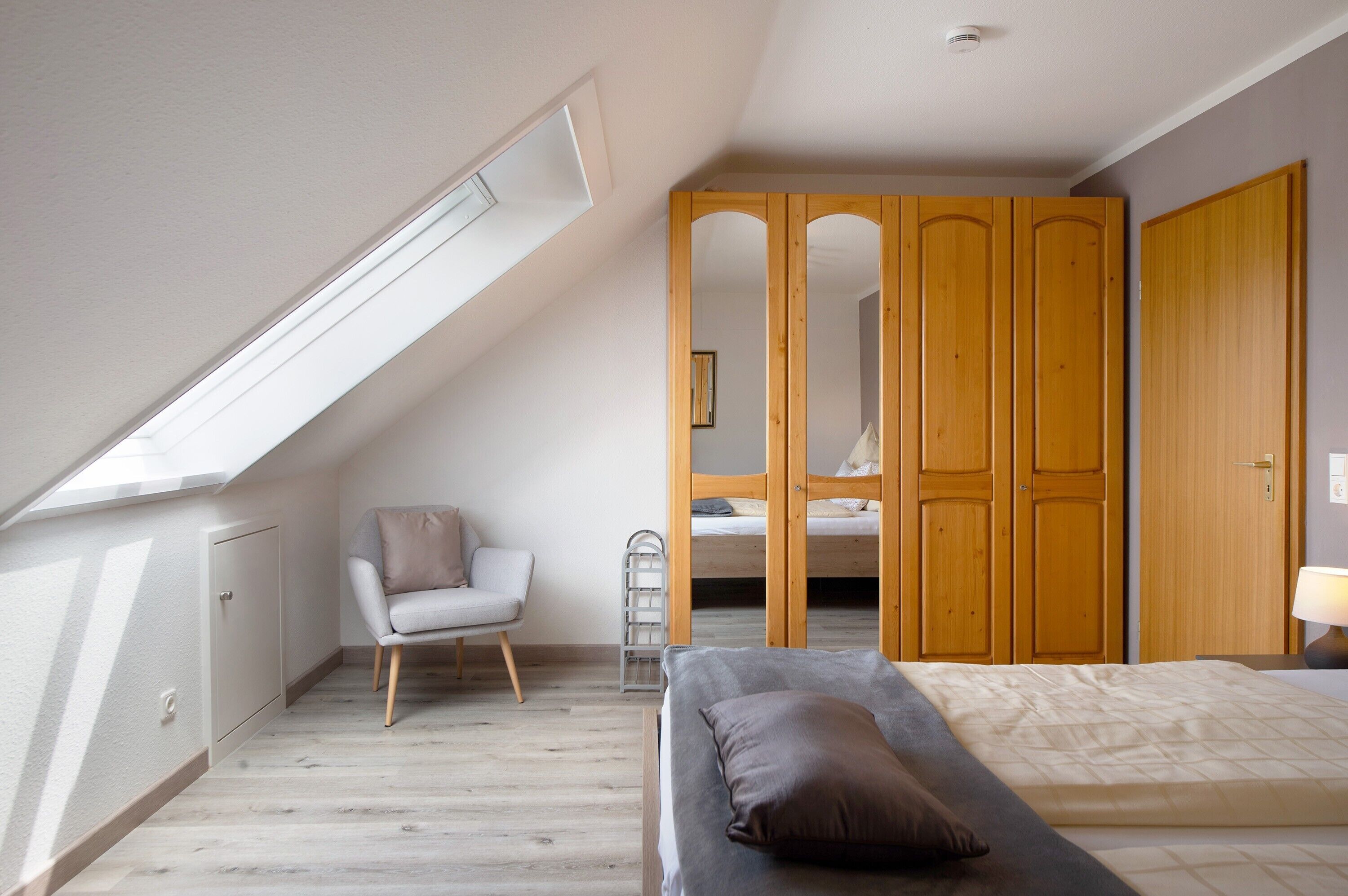 A bedroom with a sloped ceiling, a bed, a chair, a wardrobe, and a mirror.