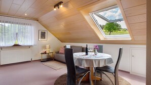 Une pièce avec un plafond en pente, un puits de lumière, une table ronde entourée de chaises, un radiateur et une fenêtre avec des rideaux.