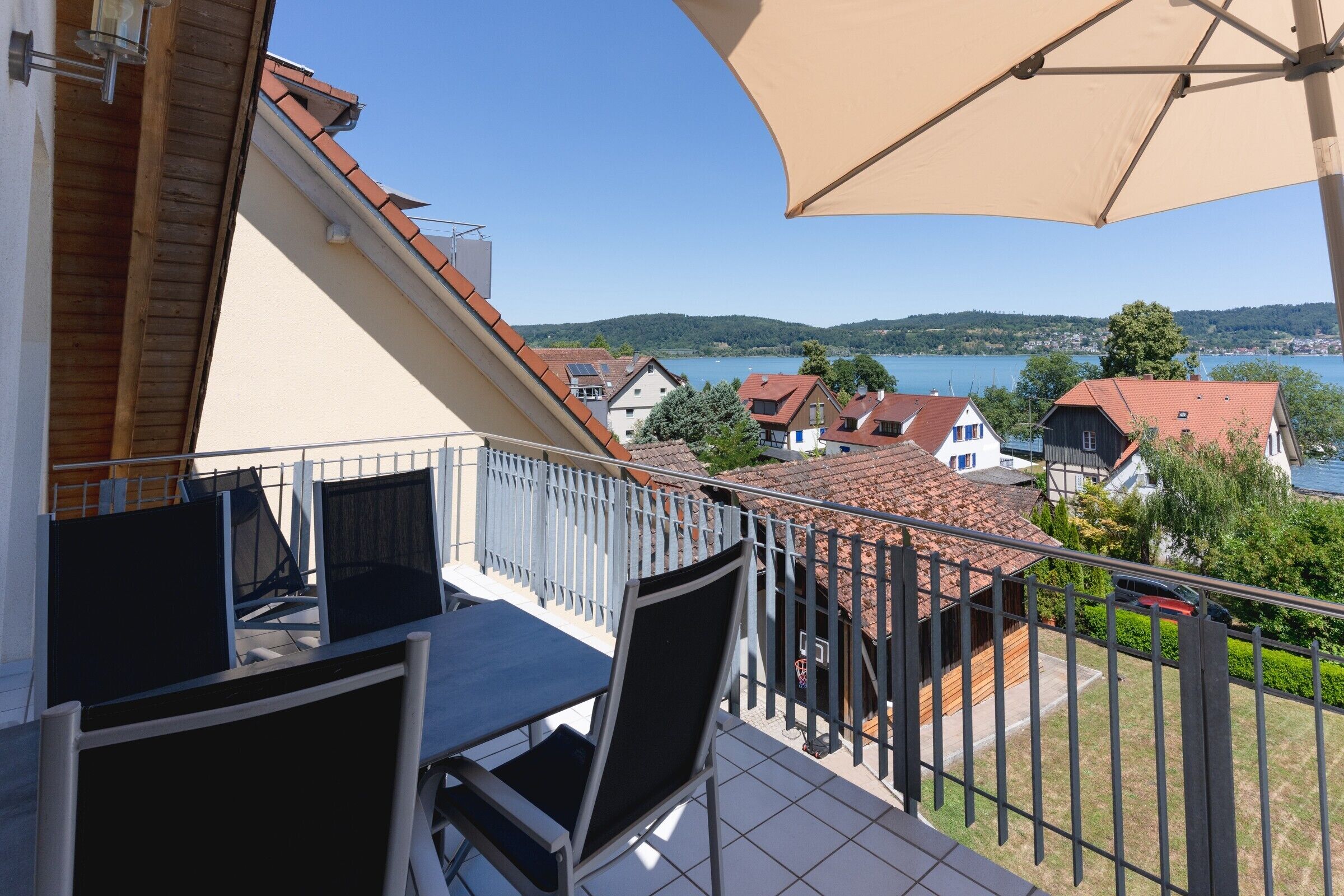 A balcony with a table and chairs, overlooking a lake with houses and a clear sky.