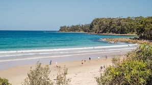 Una playa con agua azul cristalina, orilla de arena y abundante follaje verde al fondo.
