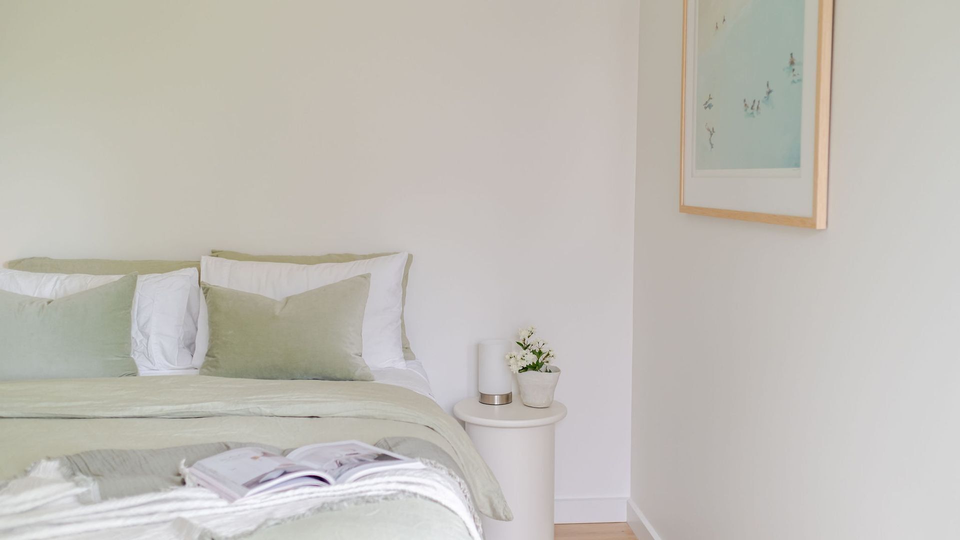 A neatly made bed with a green and white color scheme, a small bedside table with a white vase, and a framed artwork on the wall.