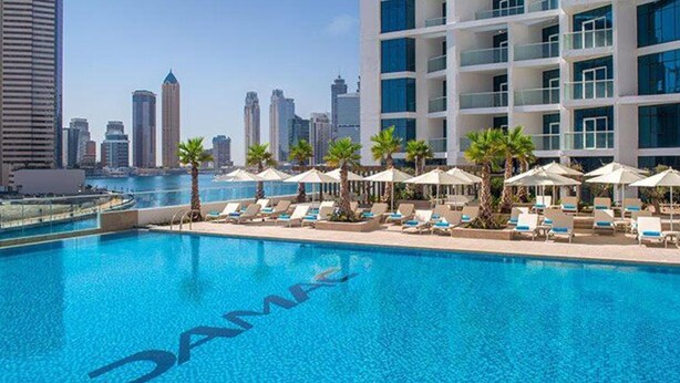 A hotel pool with lounge chairs and umbrellas, surrounded by palm trees, with a city skyline in the background. - Canal View Studio Apt - Maison Prive (Dubai)