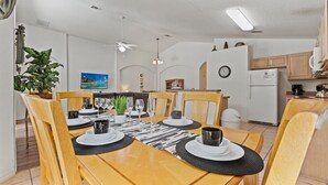 Een eethoek met een houten tafel gedekt voor vier personen, omringd door stoelen. Er is een plafondventilator, een klok aan de muur en een keuken in de achtergrond zichtbaar.