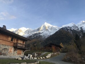 Uma cabana de madeira rústica com uma varanda, ao fundo montanhas cobertas de neve e um céu claro.