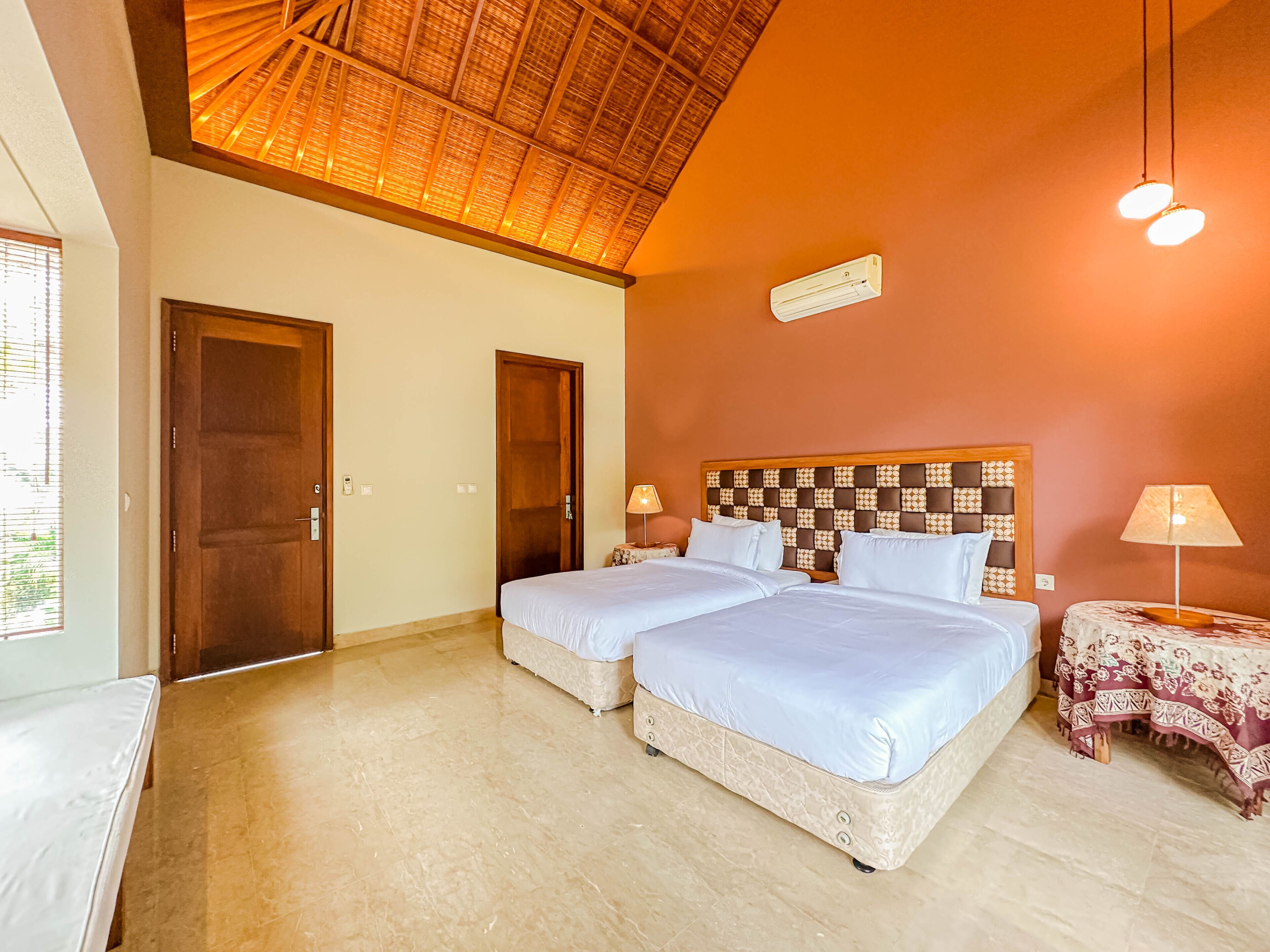 Habitación de hotel con dos camas, un ventilador de techo, aire acondicionado de pared y una mesita con lámpara.