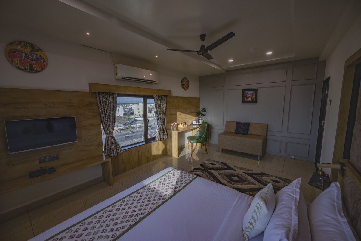 Quarto de hotel com cama, escrivaninha, cadeira, televisão, ventilador de teto e janela com cortinas.