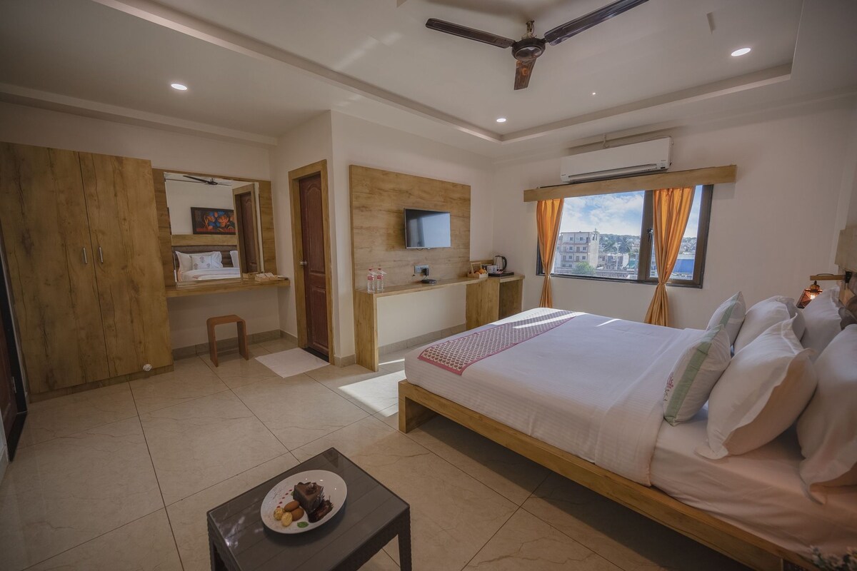 Quarto de hotel com cama, escrivaninha, cadeira, mesinha com um prato de comida, televisão e janela com cortinas.