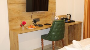 A hotel room with a wooden desk, a green upholstered chair, a flat-screen TV mounted on the wall, a small vase with flowers, a plate with oranges, a kettle, and a bed with a white duvet. - Ginger Somnath (Veraval)
