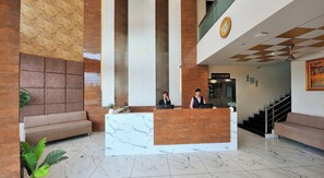 Um saguão de hotel com balcão de recepção, sofás, escada e um relógio na parede.