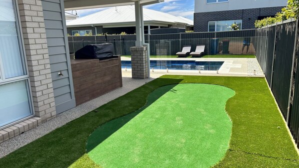 A backyard with artificial grass, a barbecue area, a swimming pool, and a fenced patio.