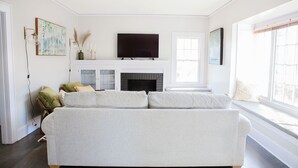A living room with a large gray sofa, a flat-screen TV above a fireplace, and a window on the right side. - Updated Campus Home (Eugene)