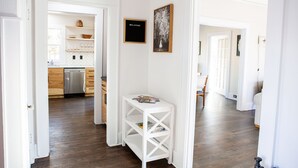 A hallway with a white door leading to a kitchen with wooden cabinets and a dishwasher. There is a white console table with a black sign that reads "WELCOME" and a framed picture on the wall. - Updated Campus Home (Eugene)