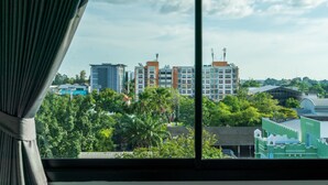 Une vue depuis une fenêtre avec des rideaux, donnant sur un paysage urbain avec des bâtiments modernes, de la verdure et un ciel dégagé.