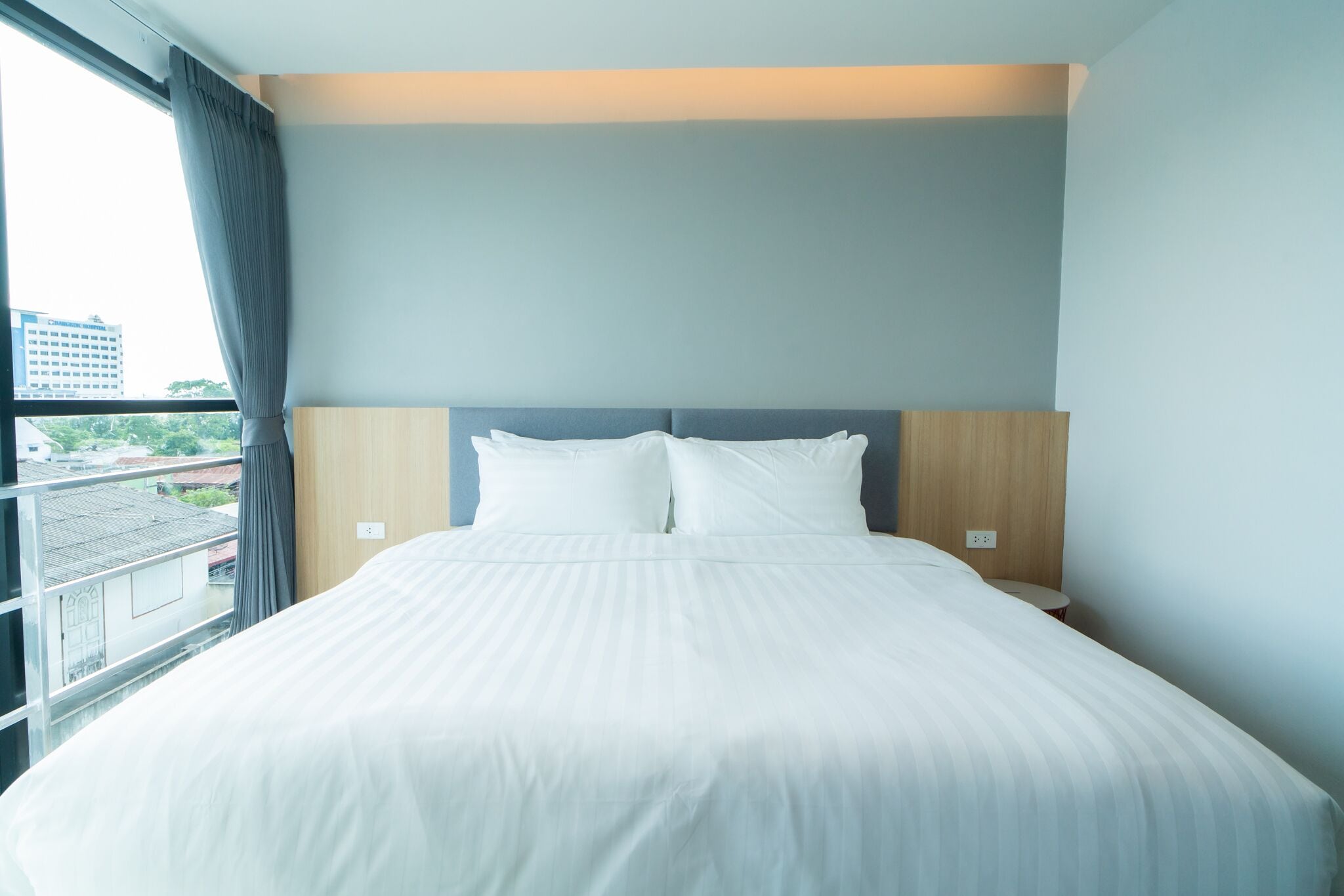A neatly made bed with white linens in a room with a view of buildings outside.
