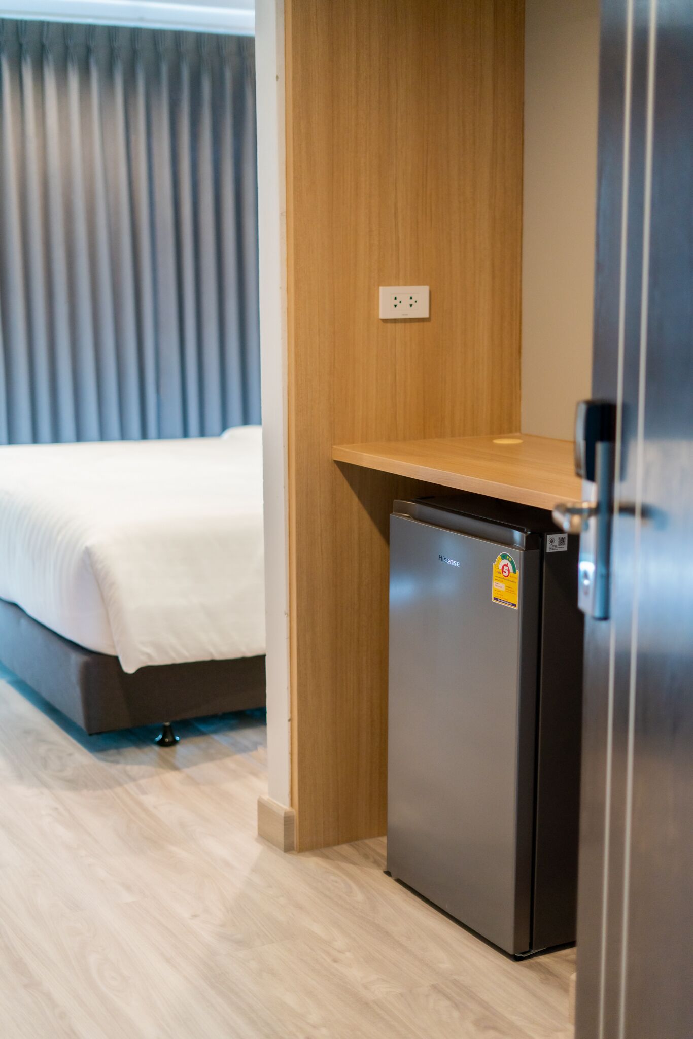 A compact hotel room with a bed, a small kitchenette with a refrigerator, and a wooden shelf.