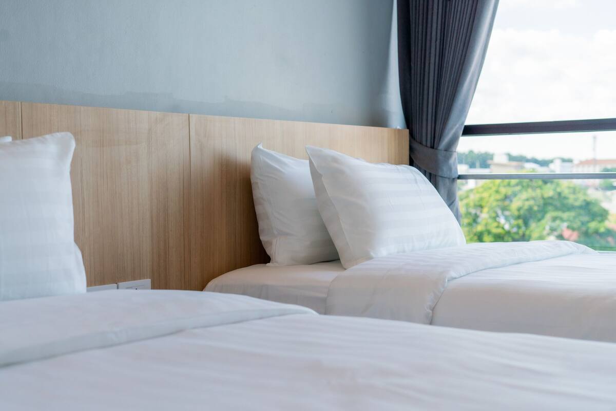 A hotel room with two beds, a wooden headboard, and a view of greenery through the window.