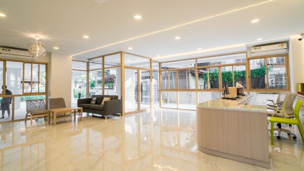 Un espace d’accueil moderne avec un comptoir de réception en bois, un coin salon avec un canapé et des fauteuils, de grandes fenêtres et un lustre.