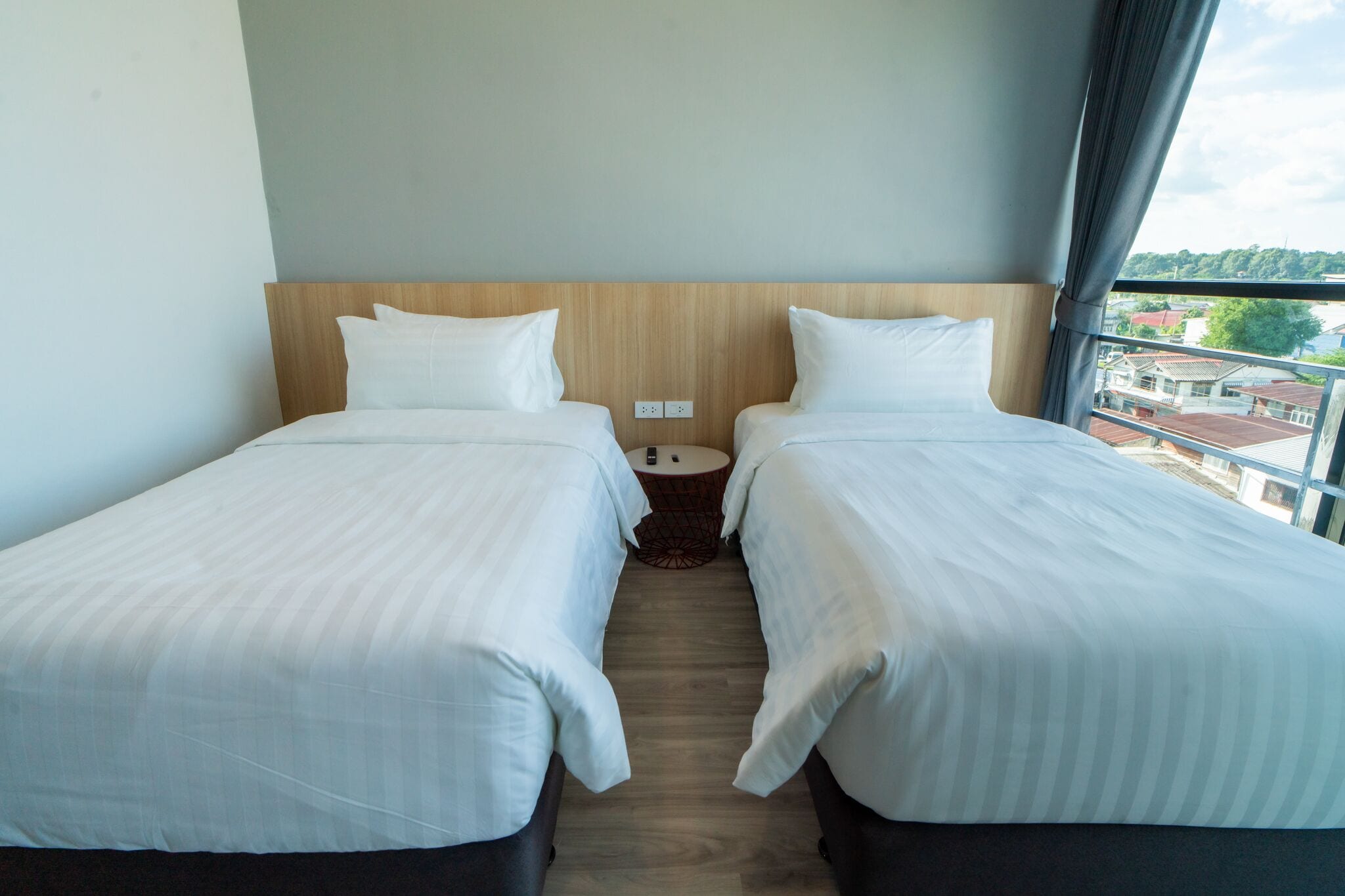 Une chambre d’hôtel avec deux lits simples, une tête de lit en bois, une petite table ronde et une vue sur la ville grâce à de grandes fenêtres.
