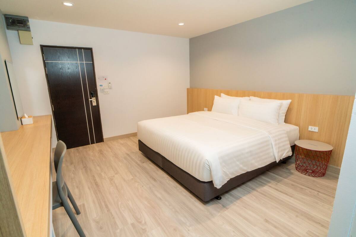 A modern hotel room with a large bed, a wooden headboard, a small round side table, and a desk with a chair.