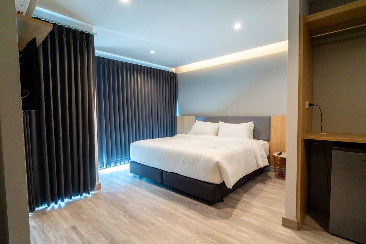 A modern hotel room with a large bed, a small bedside table, a built-in TV, and a mini-fridge.