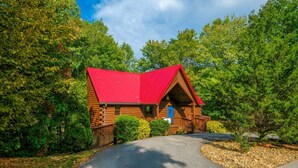 A log cabin with a red roof, surrounded by dense trees and a gravel path leading to the entrance. - Smoochin' Moose - Cozy comfort in lush surroundings! (Pigeon Forge)