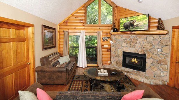 A cozy log cabin interior with a stone fireplace, a comfortable seating area, and a television mounted above the fireplace. - Smoochin' Moose - Cozy comfort in lush surroundings! (Pigeon Forge)