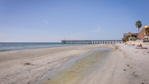 Ein Strand mit einem Steg, der sich ins Wasser erstreckt, einem sandigen Ufer und Gebäuden im Hintergrund.