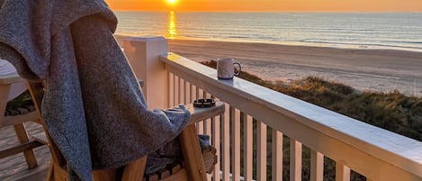 A wooden deck with a chair and a mug, overlooking a beach at sunset.