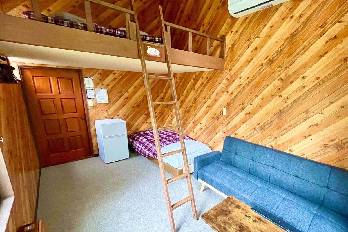 Een loft-achtige kamer met een stapelbed, een blauwe bank, een houten trap en een klein wit apparaat.