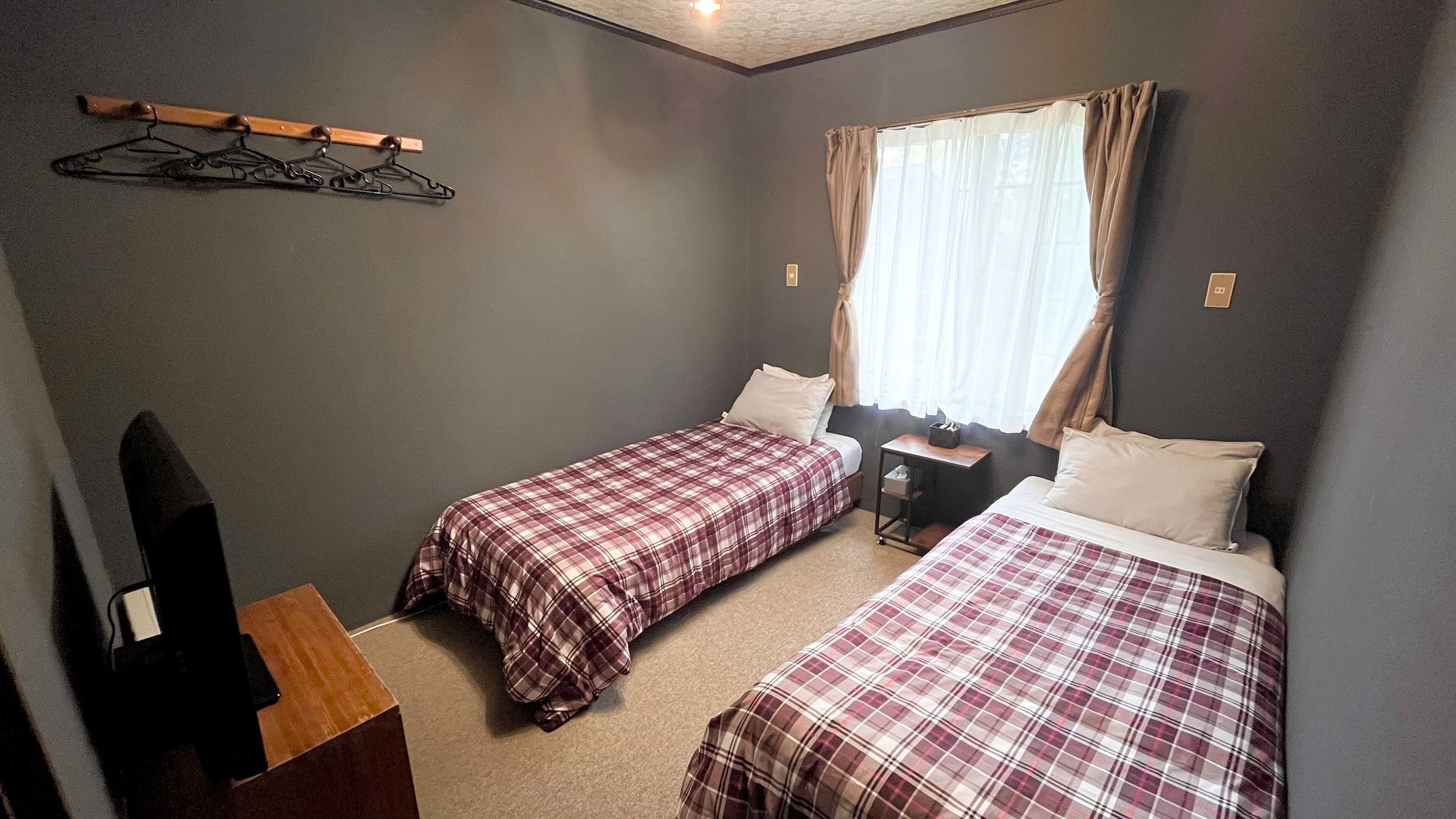 A room with two beds, a desk, a chair, a coat rack, and a window with curtains.