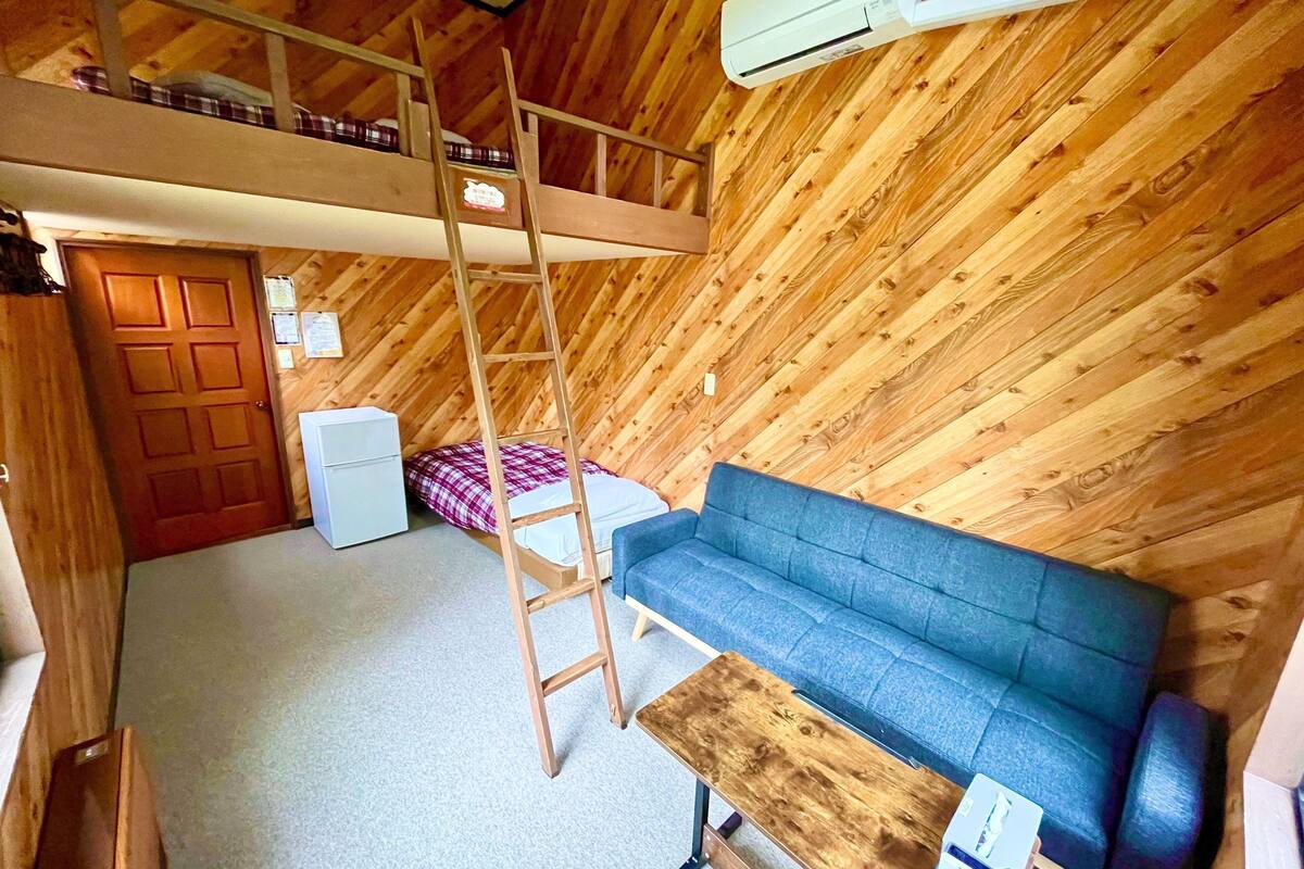 Een loft-kamer met een houten schuine plafond, een blauwe bank, een houten ladder, een kleine tafel en een witte koelkast.