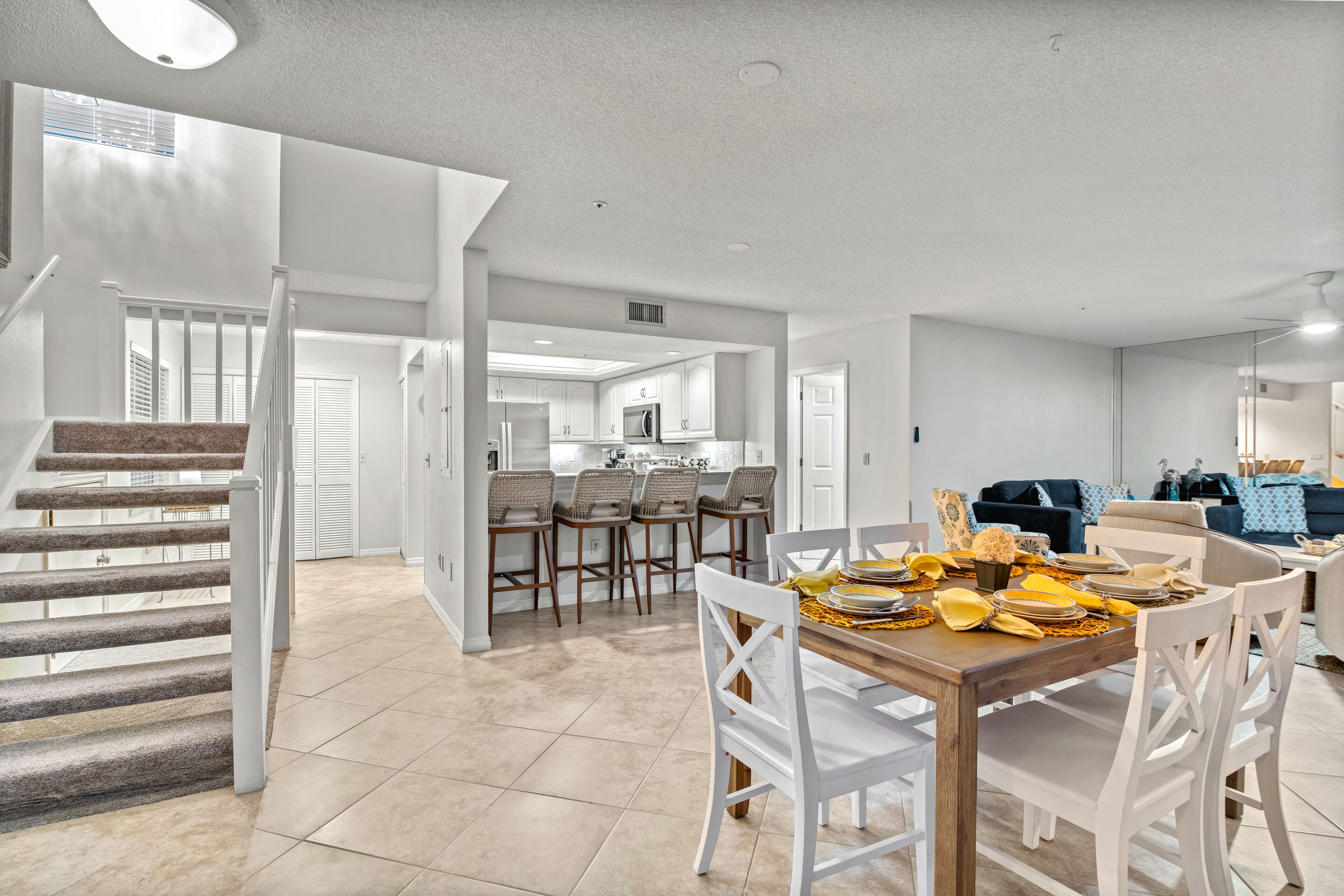 Un vaste espace de vie comprenant un coin repas avec une table dressée pour six personnes, une cuisine à l’arrière-plan et un escalier menant à un autre niveau.