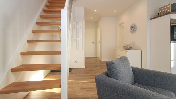 A staircase with wooden steps leading to a landing, a grey armchair, and a white cabinet with a clock on the wall. - Ferienwohnung Familienkoje (Norden)