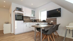 A modern kitchen with white cabinets, a built-in oven, a microwave, and a large flat-screen TV mounted on the wall. - Ferienwohnung Familienkoje (Norden)