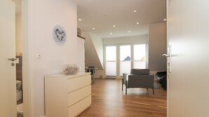 A modern interior with a wooden floor, a white dresser, a grey chair, and a large window with blinds. - Ferienwohnung Familienkoje (Norden)