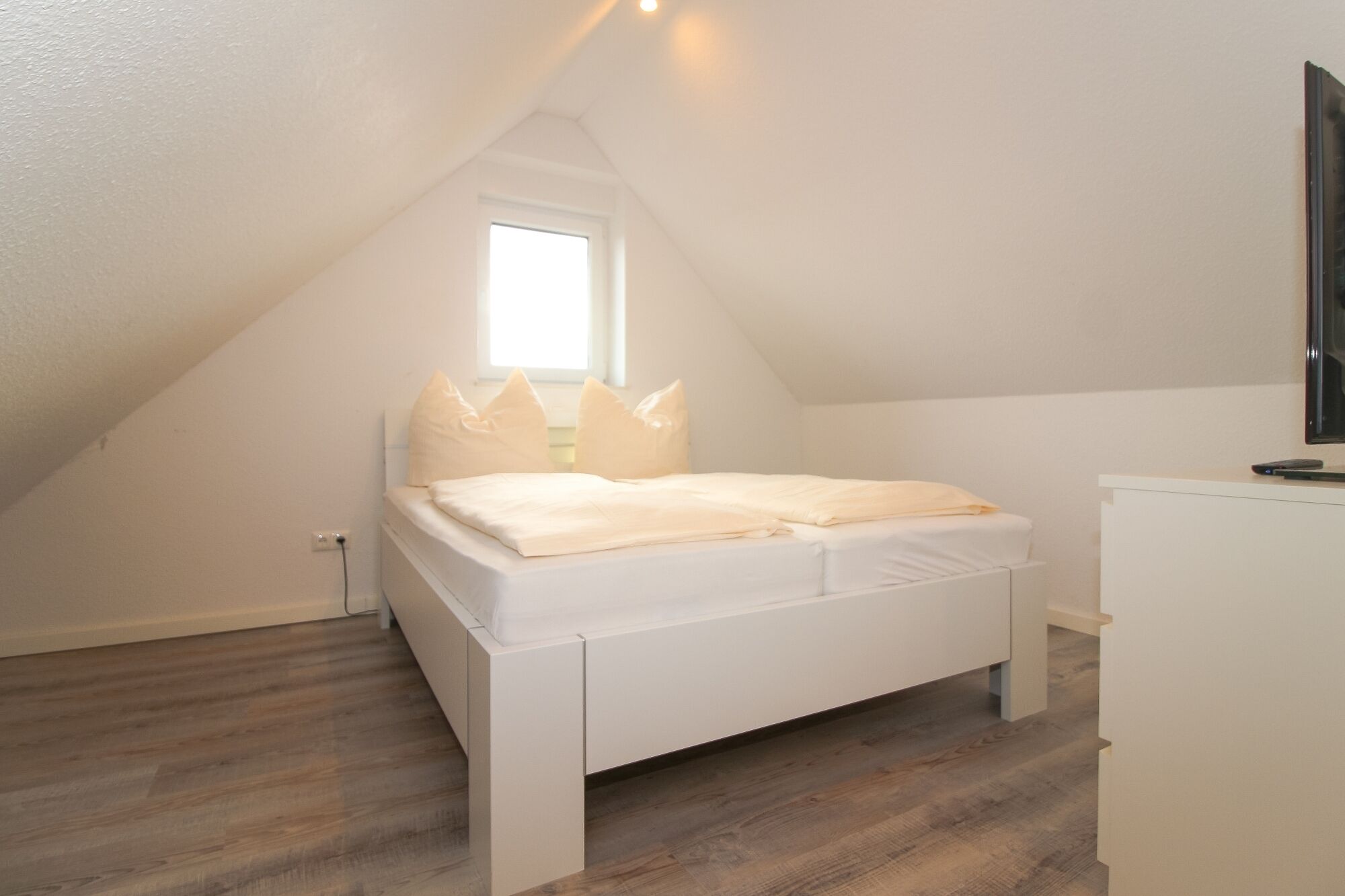 A bedroom with a sloped ceiling, a bed with white bedding, a small window, and a desk with a computer.