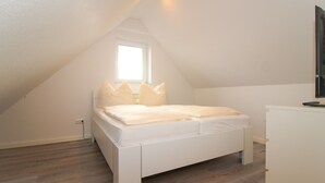 A bedroom with a sloped ceiling, a bed with white bedding, a small window, and a desk with a computer. - Ferienwohnung Familienkoje (Norden)