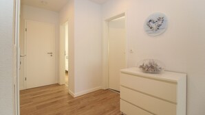 A hallway with wooden flooring, white walls, a white dresser with drawers, and a round wall clock. - Ferienwohnung Familienkoje (Norden)