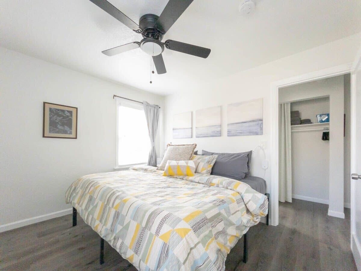 A bedroom with a bed, a ceiling fan, a window with curtains, and a door leading to another room.