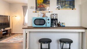 Une cuisine compacte équipée d’un four à micro-ondes, d’une machine à café et de deux tabourets. Deux tableaux sont accrochés au mur, au-dessus du comptoir.