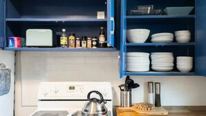 Une cuisine avec un four blanc, une bouilloire et une planche à découper en bois sur le comptoir. On y trouve des étagères remplies d’assiettes blanches et un placard bleu contenant des épices et des condiments.