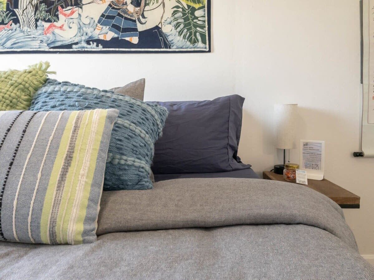 A neatly made bed with grey bedding and multiple pillows, a wall-mounted artwork depicting a traditional scene, and a small table with a lamp and a sign.