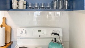 Een keuken met een wit fornuis, een blauwgroene waterkoker en een blauw kastje erboven met servies en glazen.