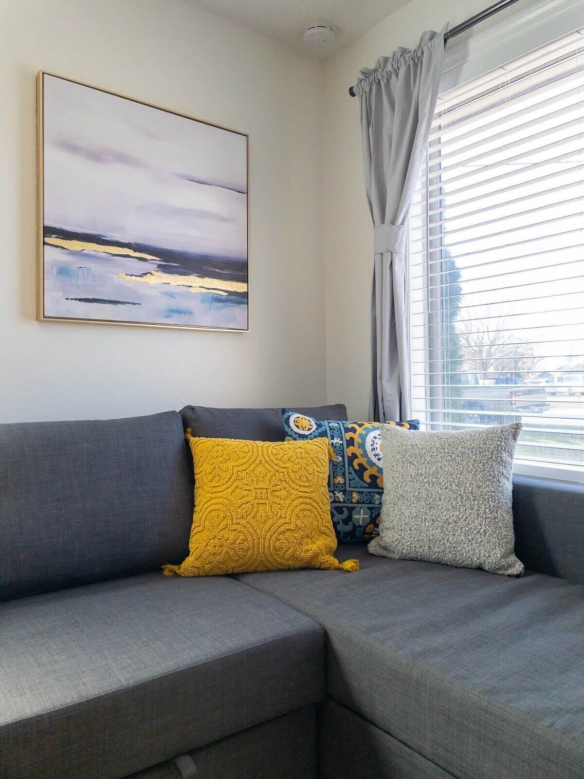A grey sofa with three patterned cushions in a room with a large window, a ceiling light, and a framed abstract painting on the wall.