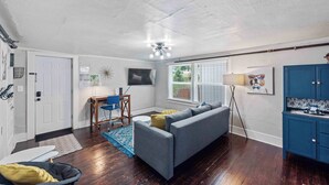 A living room with a gray sofa, a small table with chairs, a TV mounted on the wall, and a window with blinds.