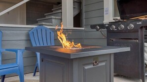 A barbecue grill with a fire burning on top, a blue chair, and a window overlooking a deck.
