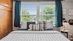 A bedroom with a bed, curtains, a bedside table, a lamp, and a view of greenery outside.