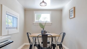 Un coin repas avec une table en bois et quatre chaises noires, une plante en pot, une fenêtre avec des stores et une citation encadrée au mur.