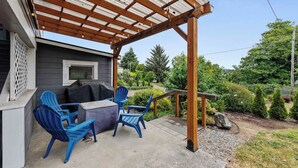 Un patio couvert avec deux chaises bleues et une table, une pergola en bois et une vue sur un jardin agrémenté d’arbres et d’arbustes.