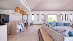 A modern living space with a kitchen area, bar stools, a sofa, and a large window providing a view of greenery.